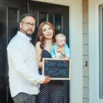 Young family with baby holding a sign 'Our First Home' at doorstep, symbolizing new beginnings.